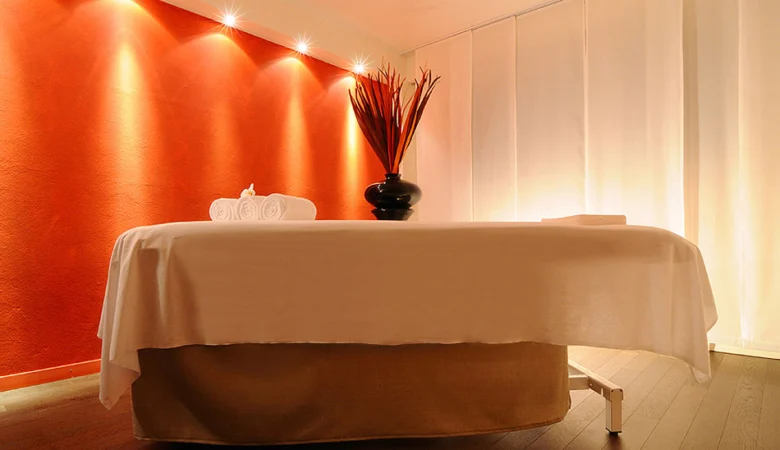 Massageliege vor einer orangen Wand im Frauenhotel Revital. Auf der Liege steht eine Pflanze und eine Schale mit Massage-Öl.